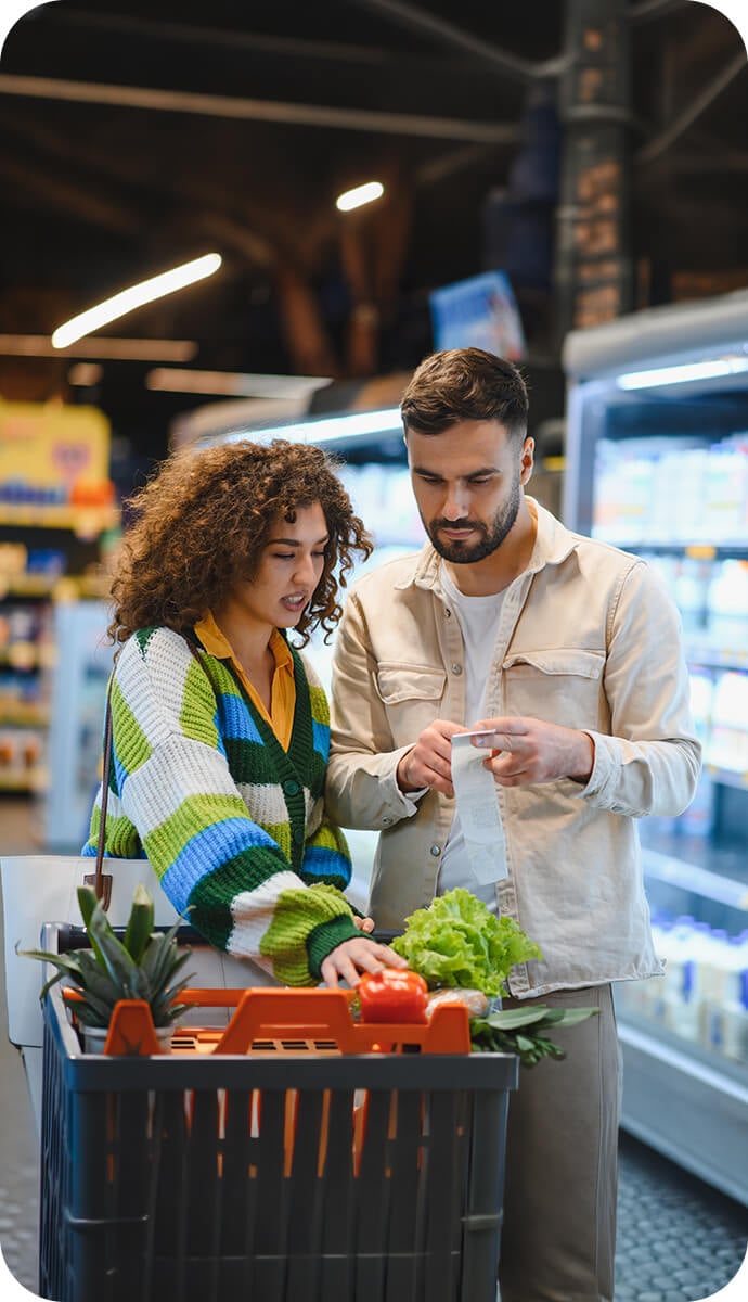 Dos personas comparan el ticket de compra junto a un carrito con alimentos en el pasillo de un supermercado. Dos personas comparan el ticket de compra junto a un carrito con alimentos en el pasillo de un supermercado.